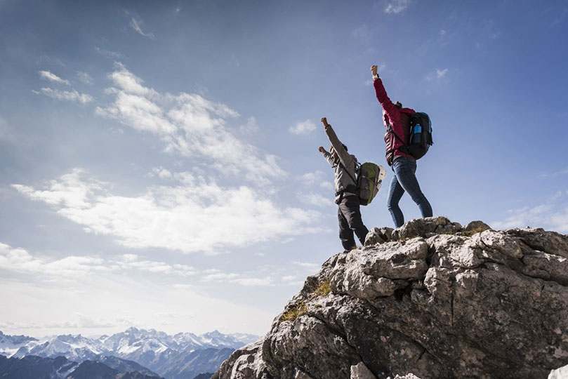 Ein Mann und eine Frau in Wanderkleidung und mit Rucksack stehen auf einem Berggipfel uns reißen freudig ihre Arme in die Höhe.