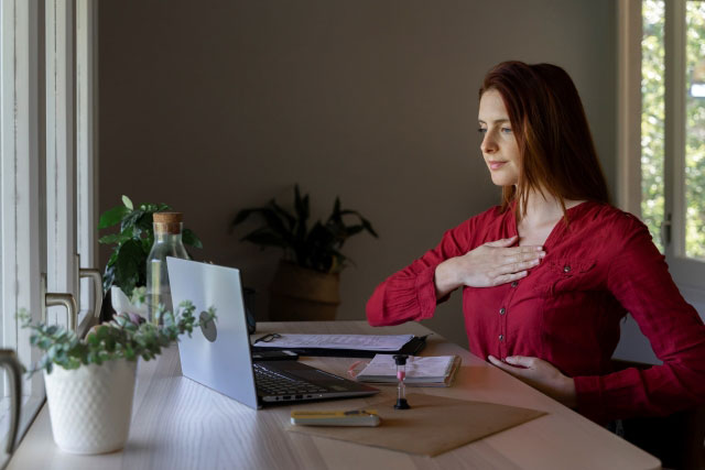 Eine Frau sitzt am Schreibtisch vor ihrem Laptop und legt ihre Handflächen auf ihren Brustkorb und ihr Zwerchfell.