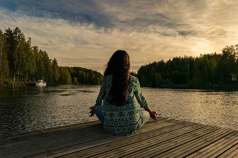 Frau sitzt mit dem Rücken zugewandt am See und macht Yoga.